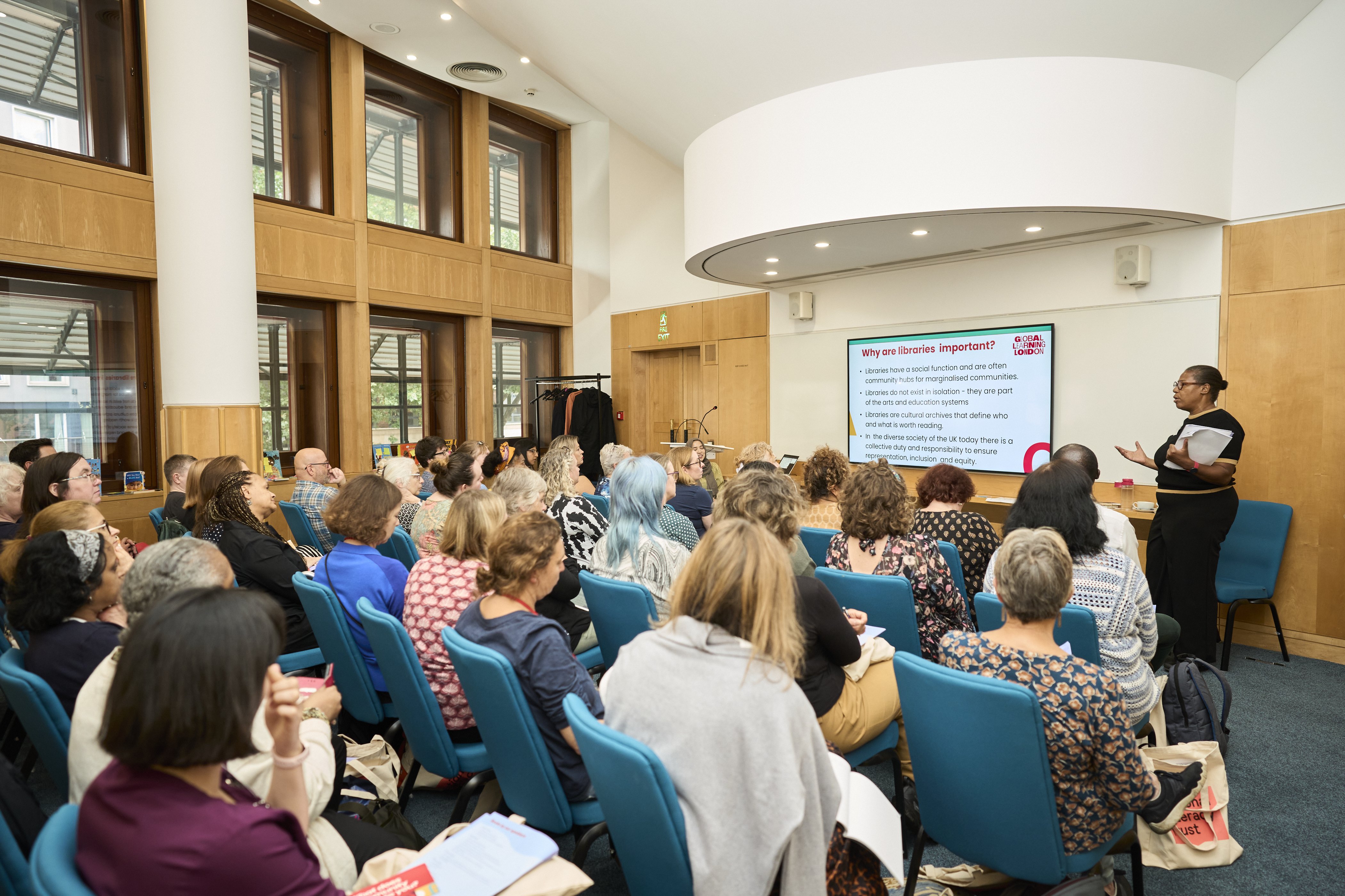 A room full of diverse people sitting on chairs, listening to a talk. The speaker is a black woman wearing black and gesturing to a powerpoint slide that reads 'why are libraries important?'