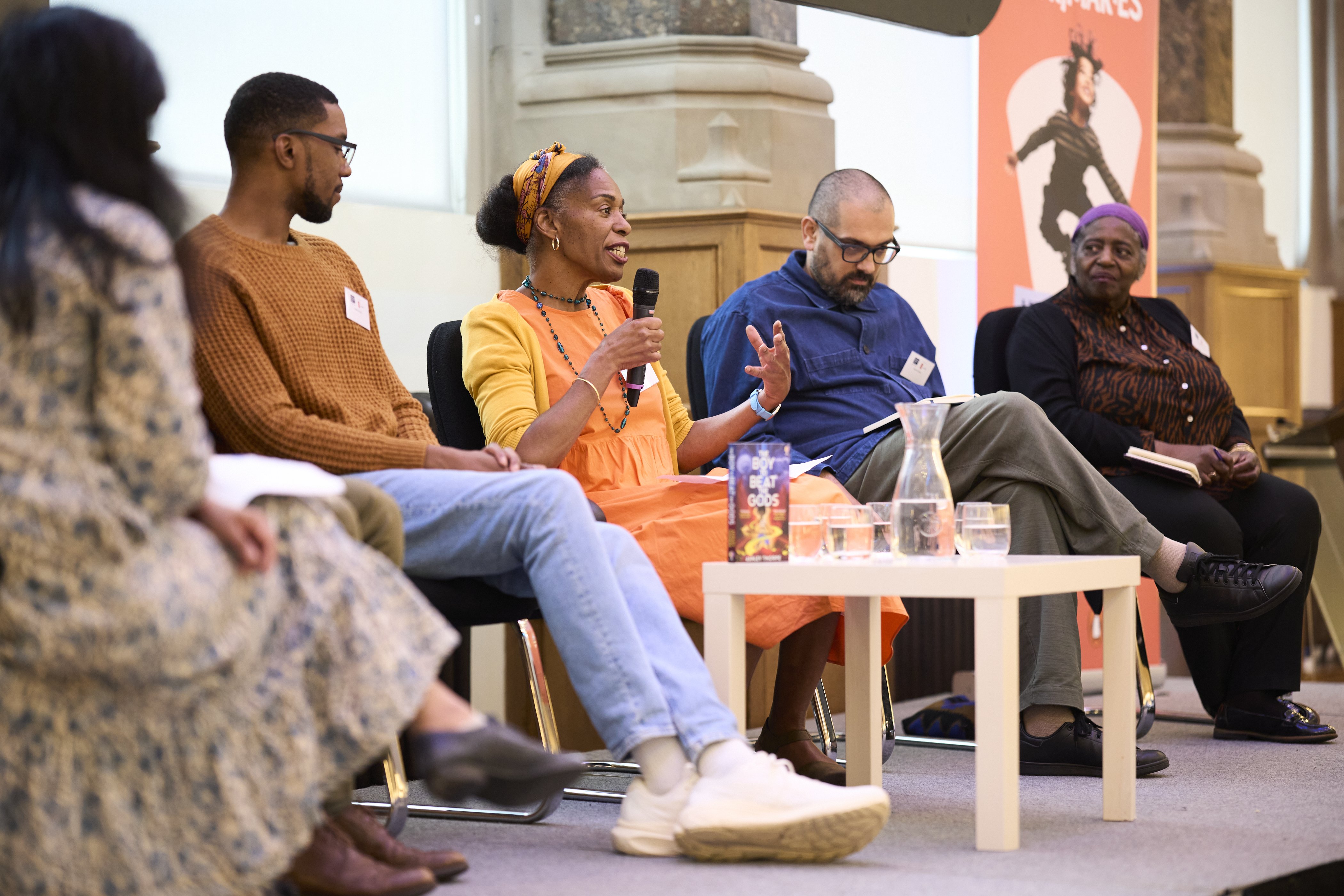 A diverse group of people in a panel discussion on a stage. A black woman in an orange dress speaks into a microphone, the others listen thoughtfully.