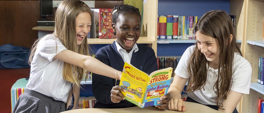 Three girls in a library wearing school uniforms look at a book together.