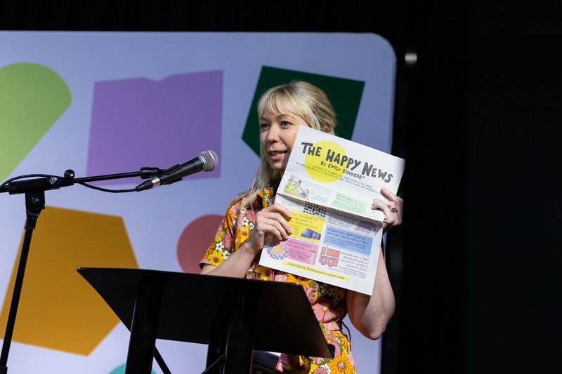 Emily Coxhead holds up a copy of The Happy Newspaper whilst talking at a lectern