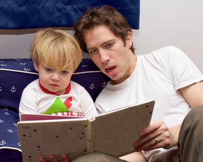 Small child and father/adult man lie on a bed reading a book together.