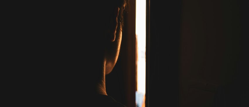 Person peers through the gap of a slightly opened door from a dark room into a light space